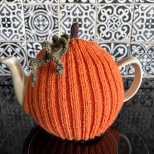 Puede incluir: Un calentador de té de calabaza naranja tejido, con un tallo marrón y detalles de vid verde. Diseñado para encajar sobre una tetera blanca, creando un tema festivo de otoño o Halloween.