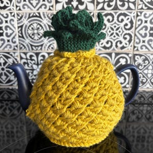 May include: A yellow crocheted tea cosy in the shape of a pineapple, with a green crocheted crown. The cosy covers a blue teapot. The yarn used to make the cosy has a textured finish.
