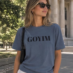 May include: A slate blue short-sleeved t-shirt featuring the word GOYIM printed in bold black capital letters across the chest. The garment has a classic crew neckline and a relaxed fit, constructed from a solid-colored fabric with a subtle texture.