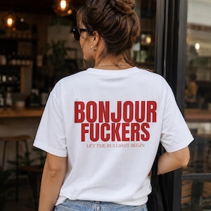 Op de afbeelding: Een wit T-shirt met korte mouwen en rode tekst op de achterkant met de tekst BONJOUR FUCKERS en daaronder kleinere rode tekst met de tekst LET THE BULLSHIT BEGIN. Het shirt wordt gecombineerd met een lichtblauwe spijkerbroek met een rood label.