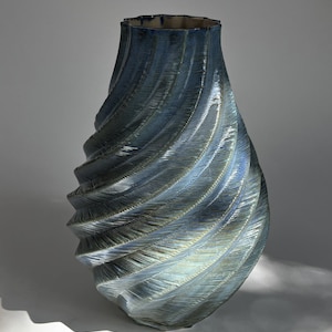May include: A ceramic vase with a deep blue and metallic finish, featuring a distinct spiral texture that wraps around the body. The vessel has a tapered neck and a wide, rounded base, with visible ridges creating a dynamic, light-reflecting surface.