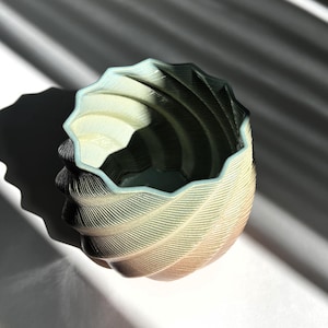 May include: A small, cylindrical planter with a spiraling, ribbed texture and a scalloped rim. The surface features a gradient color transition from a light, pale tone to a darker, muted shade, creating a distinct visual depth across the exterior.