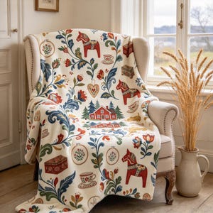 Könnte beinhalten: Eine cremefarbene Wohndecke mit einem wiederkehrenden Volkskunst-Muster aus roten Dala-Pferden, roten Häusern, blau-grünen Blumenranken, Teetassen, Holzkisten und Stickrahmen, die über einen beigefarbenen Polstersessel drapiert ist.