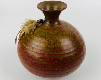 May include: A round, red and green ceramic vase with a narrow neck and a small brown bead and feathers attached to the neck.