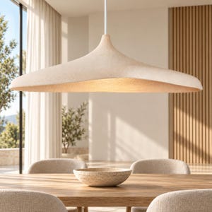 May include: A wide, saucer-shaped pendant light with a textured, off-white exterior and a warm, illuminated underside. The fixture is suspended by a single thin white cord, featuring a smooth, organic silhouette that tapers toward the center.