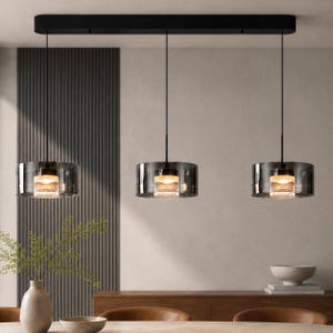 Puede incluir: Una lámpara colgante lineal con tres pantallas cilíndricas de cristal suspendidas de un dosel de techo rectangular negro. Cada pantalla consta de una capa exterior de cristal gris oscuro transparente que rodea un núcleo de cristal estriado.