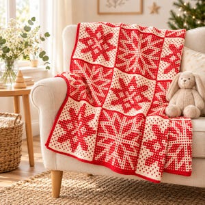 Può includere: Una coperta all'uncinetto con un motivo geometrico ripetuto di fiocchi di neve in rosso vivace e bianco crema. Il design a blocchi quadrati è realizzato con un punto testurizzato, creando un tessuto spesso e caldo con bordo rosso.