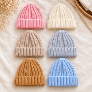 Könnte beinhalten: Eine Sammlung von sechs gerippten Strickmützen, die in zwei Spalten angeordnet sind. Die Mützen haben einen umgeschlagenen Saum und sind in den Farben Hellrosa, Creme, Beige, Hellblau, Senfgelb und einem helleren Himmelblau gehalten.