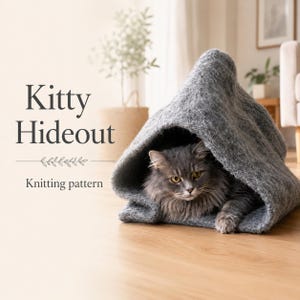 May include: A gray felted wool cat cave shaped like a tent with a long-haired gray cat resting inside. The text Kitty Hideout Knitting pattern is printed on the left side of the image in a dark gray serif font with a small decorative leaf line.
