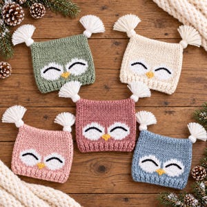 May include: Five square knitted hats featuring owl faces with white eye patches, black embroidered eyes, and yellow beaks. Each hat has two white tassels at the top corners. The hats are in sage green, cream, dusty rose, light pink, and dusty blue colors.