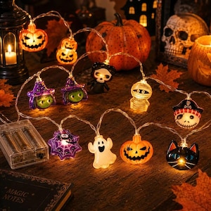 Può includere: Una catena di luci LED di Halloween con varie figure, tra cui zucche arancioni, una strega viola, un fantasma bianco, una mummia, un gatto nero, una ragnatela e un teschio, collegate da un filo trasparente e alimentate da un vano batterie.