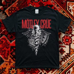 以下が含まれることがあります： 赤い文字でMÖTLEY CRÜEと書かれ、その下に杖に巻き付いた翼のある蛇の白いグラフィックが描かれた黒い半袖Tシャツ。デザインには円形の背景要素が含まれており、標準的なクルーネックのコットンTシャツにプリントされています。