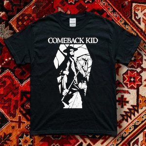 May include: A black short-sleeved t-shirt featuring a white graphic print of a figure in a hat holding a long tool inside a coffin-shaped outline. The text COMEBACK KID is printed in white capital letters above the graphic on the chest area.