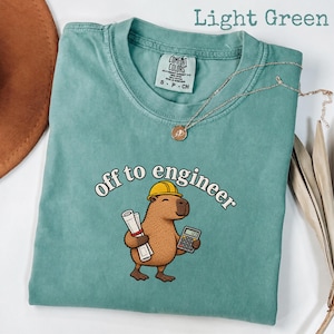 Op de afbeelding: Een lichtgroen t-shirt met een afbeelding van een bruine capibara met een gele veiligheidshelm, die een opgerolde blauwdruk en een rekenmachine vasthoudt. De tekst "off to engineer" staat in een wit gebogen lettertype boven de capibara.
