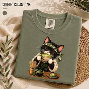 May include: A moss green Comfort Colors 1717 t-shirt featuring a graphic of a black and white cat wearing a green headband and apron, holding a bowl of green matcha tea. A bamboo whisk and a small bowl of matcha powder are on a wooden table.