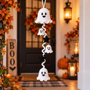 May include: A vertical hanging decoration featuring three white crocheted ghost figures and two black crocheted stars arranged along a central white spiral cord. Each ghost has two black embroidered eyes and a black embroidered mouth.