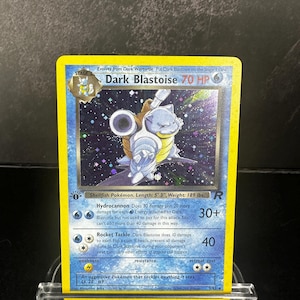 May include: A Dark Blastoise trading card featuring a blue Pokémon with cannons on its back. The card has a yellow border, a holographic background with stars, and text including 70 HP, Hydrocannon, and Rocket Tackle attacks. It is a stage 2 card.