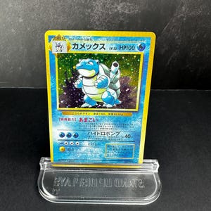 May include: A Japanese Blastoise trading card featuring a holographic illustration of the blue turtle-like creature. The card displays the name Kamex, level 52, 100 HP, and water-type symbols. It is held upright in a clear plastic display stand.