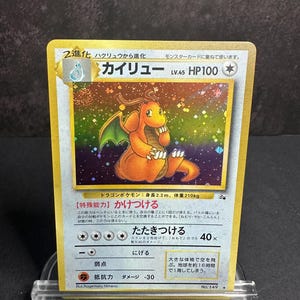 May include: A Japanese Dragonite Pokémon trading card featuring a holographic rainbow background. The card displays the character illustration, text in Japanese, HP100, and level 45. The border is gold, and the card is set in a clear plastic stand.