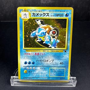 May include: A Japanese Blastoise trading card featuring a holographic illustration of the blue turtle creature with cannons on its back. The card displays the name Kamex, 100 HP, and various game statistics in Japanese text against a yellow border.
