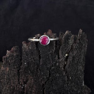 Peut inclure: Une bague en argent avec une pierre précieuse ronde, facettée et d'un rouge rosé profond, montée dans un serti clos lisse. L'anneau est en argent poli et la pierre présente une surface texturée à multiples facettes.