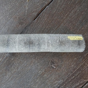 May include: A cylindrical gray stone specimen with a rough, textured surface. A small yellow rectangular label is affixed to the right side, featuring the printed text "Nodite" and "Alaska" in black ink, indicating the origin of this geological sample.