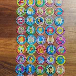 Könnte beinhalten: Eine Sammlung von fünfzig runden Pogs aus Pappe mit farbenfrohen Illustrationen verschiedener Charaktere aus den Simpsons. Jede Scheibe zeigt ein Charakterporträt, den Namen in Fettdruck und eine einzigartige Hintergrundfarbe, angeordnet in einem Raster.