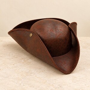 May include: A dark brown tricorn hat with a weathered, textured finish. The hat features a rounded crown and a wide, upturned brim that is secured on one side with a small, circular brass-colored metal stud, creating a classic three-cornered shape.