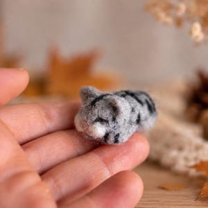 Puede incluir: Una pequeña figura de gato atigrado gris y blanco hecha con fieltro de aguja en posición de dormir. El gato en miniatura presenta rayas gris oscuro sobre su cuerpo suave y difuso, orejas puntiagudas y un hocico blanco de lana.