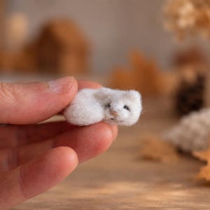 Puede incluir: Una figura de animal en miniatura hecha de lana fieltrada blanca con una textura suave y esponjosa, ojos pequeños y oscuros y una nariz diminuta. La pequeña escultura artesanal tiene forma de criatura acurrucada y se sostiene entre los dedos.