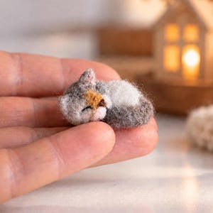 Puede incluir: Una figura de gato en miniatura hecha de fieltro de aguja en posición de descanso. El gato presenta una mezcla de parches de pelaje blanco, gris y marrón claro, con orejas pequeñas y puntiagudas y una textura suave y esponjosa de lana.