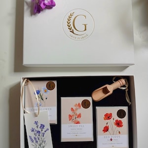 Könnte beinhalten: Eine weiße Geschenkbox mit goldenem Grow in Grace-Logo und violetten Blumen darauf. Im Inneren befinden sich drei Samentütchen mit den Aufschriften Cornflower, Sweet Pea und Red Poppy, eine kleine Holzschaufel und ein Blumenanhänger.