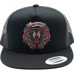 May include: A black trucker cap with a mesh back and a flat peak. The front features an embroidered graphic of a roaring bear surrounded by red roses and two crossed arrows. Above the bear, a banner contains the text WILD RIDE in black letters.