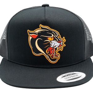 Może przedstawiać: Czarna czapka typu trucker snapback z grafiką głowy warczącej pantery w kolorach czarnym, białym, pomarańczowym i czerwonym. Czapka posiada sztywny przedni panel, siateczkowe panele boczne i tylne oraz płaski daszek z okrągłą naklejką po prawej stronie.