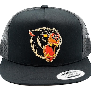 May include: A black snapback trucker cap featuring a mesh back and a flat brim. The front panel displays a graphic illustration of a snarling bear head with yellow eyes, orange and red accents, and sharp teeth, with a circular sticker on the brim.