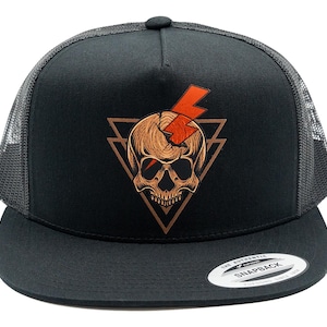 Könnte beinhalten: Eine schwarze Trucker-Cap mit Netzrückseite und flachem Schirm. Auf der Vorderseite befindet sich eine Grafik eines beigen Schädels mit gesprungener Stirn, einem leuchtend orangefarbenen Blitz und zwei überlappenden braunen Dreiecken. Ein Aufkleber befindet sich auf dem Schirm.