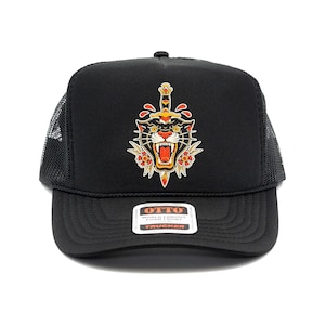 Puede incluir: Una gorra trucker negra con un gráfico de una cabeza de pantera negra rugiente atravesada por una daga vertical, decorada con flores rojas y amarillas. La pegatina en la visera dice OTTO WORLD FAMOUS FOAM FRONT TRUCKER.