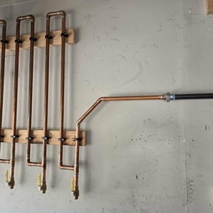 May include: A wall-mounted plumbing system featuring four vertical copper pipe loops secured to two horizontal wooden boards. Each loop includes a yellow-handled ball valve at the bottom. A copper pipe extends to the right, connecting to a black iron pipe.