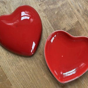 May include: A glossy red heart-shaped trinket box with a removable lid. The object features a smooth, reflective finish on both the convex lid and the concave base, showcasing a vibrant solid red color throughout the entire heart-shaped structure.