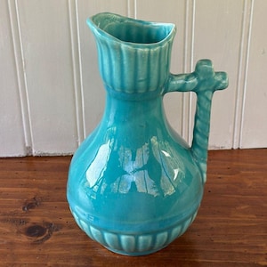 May include: A turquoise ceramic pitcher with a glossy finish, featuring a bulbous body, a flared rim with vertical fluting, and a sturdy handle with a twisted rope-like texture that extends slightly above the top edge of the vessel.