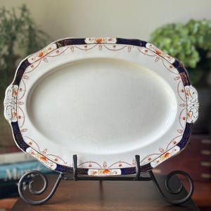 May include: An oval ceramic serving platter featuring a cream-colored center and a decorative border. The rim is adorned with a deep navy blue band, intricate orange floral accents, and delicate gold-toned filigree patterns on a white background.