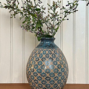 May include: A rounded ceramic vase with a narrow neck, featuring a teal and tan geometric floral pattern across its body. The vase has a mottled teal finish at the top and contains a bouquet of small purple wildflowers with green stems.
