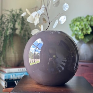 May include: A spherical, glossy, dark mauve ceramic vase with a smooth, reflective surface. The vase features a small circular opening at the top, currently holding a delicate, thin-stemmed plant with translucent, coin-shaped white leaves.