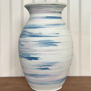 May include: A ceramic vase with a flared rim and a rounded body, featuring a white base color decorated with horizontal, streaky brushstroke patterns in shades of blue and soft pink, creating a textured, layered appearance.