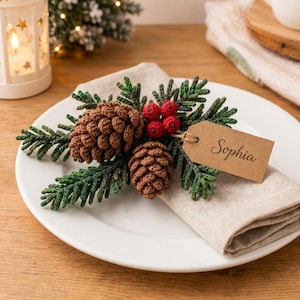May include: A crochet holiday place setting decoration featuring two brown pinecones, green pine needles, and three red berries. A rectangular tan paper tag is attached with twine, featuring the name Sophia printed in black cursive script.