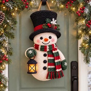 May include: A crocheted snowman wall hanging featuring a black top hat with a red band, green and red striped scarf, and three black buttons. The snowman wears green mittens and holds a small, lit black lantern. It is suspended by a rope.