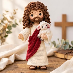 May include: A crochet doll depicting a figure with long brown curly hair, a beard, and a crown of thorns. The figure wears a cream-coloured robe with a red sash and holds a small white lamb. The doll has black eyes and brown sandals.