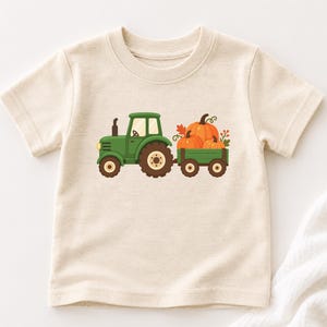 Puede incluir: Una camiseta de manga corta de color crema con un estampado gráfico de un tractor verde que tira de un pequeño remolque verde lleno de tres calabazas naranjas, enredaderas verdes y hojas de colores otoñales en el frente.