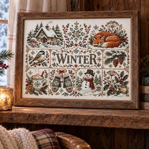 Puede incluir: Un bordado en punto de cruz enmarcado que presenta la palabra WINTER en el centro, rodeada de ilustraciones estacionales que incluyen una cabaña nevada, un zorro durmiendo, un pájaro, una taza de cacao, manoplas y un muñeco de nieve en un marco de madera.