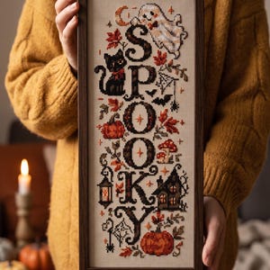 May include: A rectangular framed cross-stitch sampler featuring the vertical text SPOOKY in black letters. The design includes a white ghost, a black cat, orange pumpkins, red autumn leaves, a small house, a lantern, and mushrooms on a beige fabric.